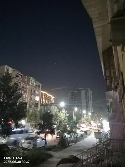 The image is a nighttime street scene in the city of Bokhtar, Tajikistan. On the left, a multi-story apartment building with numerous windows and air conditioning units is visible, partially obscured by trees. Further down the street, another building is illuminated with a string of warm lights. In the background, a tall, modern building under construction stands out. The street is lined with trees and parked cars. A white sedan is parked in the foreground. In the foreground on the right, a building facade with windows and a metal railing leading up stairs is visible. The sky is dark, with a few faint stars visible. The timestamp "OPPO A54 2026/06/30 20:37" is overlaid on the bottom left corner of the image.