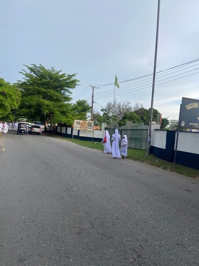 The image shows a street scene in Calabar, Nigeria. A group of people in white robes are walking on the right side of the road, near the Naval War College of Nigeria. A sign for "Eastern High School" is also visible. A green Nigerian flag is flying in the background. A row of parked cars lines the left side of the road, under a canopy of trees. Overhead power lines run across the sky. The road is paved with asphalt. The sky is overcast.