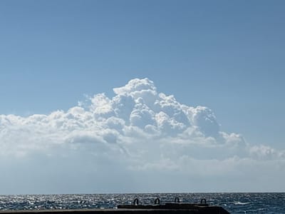 The image depicts a bright, sunny day over a body of water, likely the Black Sea, with a large, cumulus cloud formation dominating the sky. The water is a deep blue and shows a ripple effect from the wind, with sunlight glinting off its surface. In the foreground, there is a concrete structure partially submerged in the water, with visible metal loops protruding from its surface. These loops appear to be part of a pier or docking facility. The sky is a clear, light blue, with the exception of the massive cloud bank which has a mountainous, textured appearance. The overall scene suggests a coastal environment on a clear day, possibly in the vicinity of Кореизский поселковый совет, Ukraine, given the context provided. There are no people or animals visible in the frame.