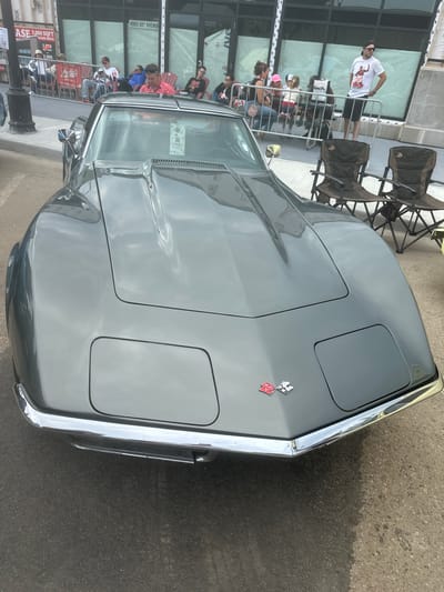 An exterior, eye-level, medium shot captures a grey Chevrolet Corvette parked on asphalt, facing forward. The car’s hood slopes down to a chrome bumper. Two checkered flags with red and white colors are affixed to the hood, below the windshield. In the background, several people are seated and standing near a building with large windows. Some individuals are sitting on chairs and others are standing in front of a black metal fence. The building has signage that reads "4905 50TH AVENUE". To the left of the Corvette, a black lamppost is visible. Two brown folding chairs are set up in front of the building, to the right of the car. The scene appears to be set in a city environment, possibly during an outdoor event or car show.