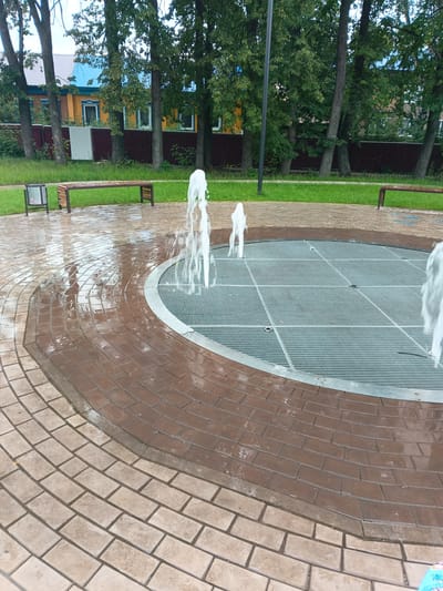 The image captures an outdoor public space in the city of Tolbazy, Russia, featuring a circular dry fountain. The fountain's surface is a metal grate with a grid pattern, surrounded by brick paving. Water jets are actively spraying upwards from the grate, creating a dynamic visual element. The paving stones are wet, indicating recent activity or rainfall. In the background, several trees with lush green foliage are visible, with a wooden fence and a multi-colored residential building behind them. A park bench and a trash receptacle are situated on the grass beyond the paved area, suggesting a recreational setting. The overall scene is one of a well-maintained urban park or public square.