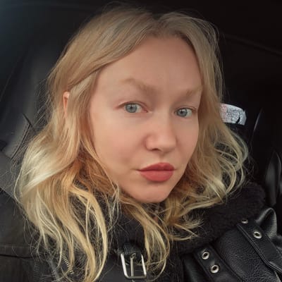 The image shows a woman with blonde hair, blue eyes, and pink lipstick. She is wearing a black leather jacket with a silver buckle and grommets. The background is dark, possibly the interior of a car. There is no information to determine if the image was taken in the city of Skotnoe, Russia.