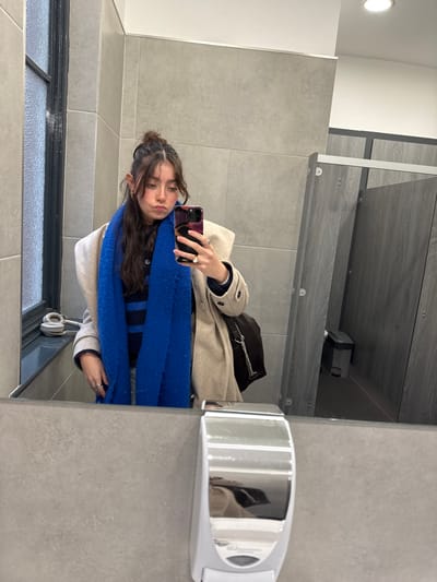 A woman is seen in a mirror selfie, wearing a tan coat, a striped shirt, and a vibrant blue scarf. She holds a phone with a pink case. A black purse sits on top of the coat. Headphones are visible near the bottom edge of the mirror. The reflection shows a bathroom setting with gray tiled walls and metal fixtures. The scene is brightly lit by an overhead light.