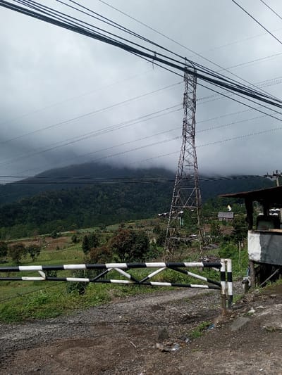 The scene, located in Saing Ta Mawt Lay, Myanmar, presents a mountainous rural landscape under a heavily overcast sky with low-hanging clouds partially obscuring distant peaks. In the immediate foreground, a black and white striped vehicular barrier is positioned across a dirt and gravel road. To the right of the barrier, a small, elevated utility or guard structure with a dark roof and white lower facade is partially visible. A prominent, tall, metallic lattice-structure electricity transmission tower stands in the midground, extending into the upper frame. Multiple overhead power lines are strung across the sky, connecting to this tower and another pole on the far right. The midground also features cultivated fields and sparse vegetation on sloping terrain, interspersed with scattered trees and distant small buildings. No individuals are present, and no active interactions or specific actions are depicted.