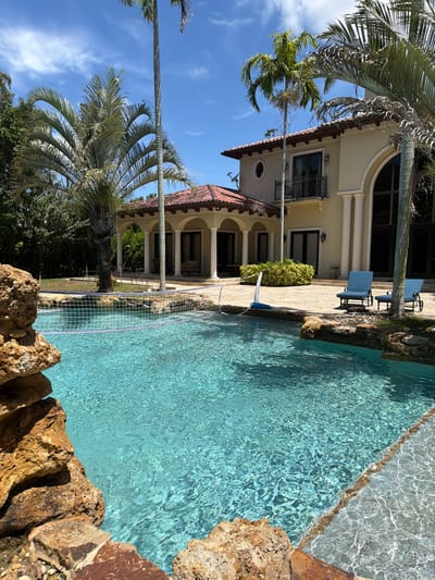 In Pinecrest, Florida, a luxurious resort-style swimming pool with clear turquoise water is a focal point. A volleyball net is stretched across a section of the pool, suggesting recreational activity. The pool is framed by large, natural-looking rocks and a stone-tiled deck. Adjacent to the pool is a grand, two-story house with a Mediterranean architectural style, featuring a red tile roof, arched doorways, and large windows. Several tall palm trees surround the property, casting shadows on the sunny, blue-sky day. Two blue lounge chairs sit on the patio near the house, overlooking the pool.