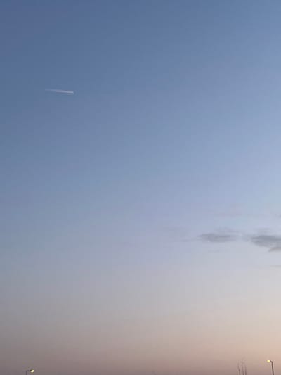 An outdoor image captures a broad expanse of sky over Roodt-sur-Syre, Luxembourg, during what appears to be twilight. The sky exhibits a gradient, transitioning from light blue at the top to a pale pinkish-orange towards the horizon. A single, thin, white linear contrail is visible in the upper left quadrant of the frame, indicative of an aircraft's passage. In the lower right quadrant, a small cluster of diffuse, light grey clouds is present. The extreme bottom edge of the image reveals a dark, indistinct silhouette of the horizon, potentially showing parts of the landscape or distant structures, including a partial view of a lit street lamp on the far left. No individuals, explicit actions, or direct interactions are discernible within the frame.