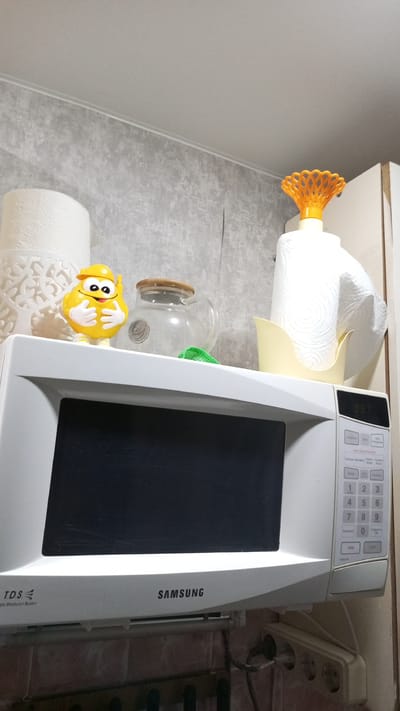 The image shows a close-up, slightly upward-angled view of the top of a white Samsung microwave oven. On top of the microwave, from left to right, are two rolls of toilet paper, one patterned and the other plain, a small yellow toy figurine with a hard hat and gloves, a glass jar with a wooden lid filled with a clear liquid, and a holder with a roll of paper towels. The microwave has a black screen and a white control panel with buttons and a digital display showing "957". The wall behind the microwave has a textured gray wallpaper. Below the microwave, a power strip with plugs is visible. The scene appears to be in a kitchen. There is no visible indication of the specific location in Sayansk, Russia.