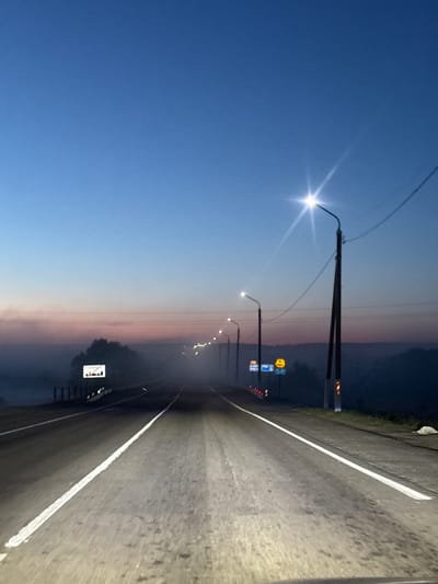 The image depicts a highway at dusk or dawn, possibly near Krychau, Belarus, based on the Cyrillic script on the road sign. A row of streetlights illuminates the road, stretching into the distance. The sky transitions from a deep blue to a light pink/orange hue on the horizon. A white line marks the edge of the road, and there appears to be some vegetation on the shoulder. Several road signs are visible, including one that may be an "end of speed limit" sign. The road surface is dark, suggesting recent rain or dampness.