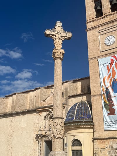 Here is a detailed description of the image:
**Scene:** An outdoor, daytime view focusing on the "Sant Lluis Beltran Cross" (Creu de Sant Lluís Beltrán) in Torrent, Spain.
**Subjects and Objects:**
* **The Cross:** An ornate stone cross atop a tall, fluted column. The cross is intricately carved.
* **Building:** The "Iglesia de la Asunción de Nuestra Señora" (Church of the Assumption of Our Lady). Parts of the church are visible in the background, including its tiled dome (blue and white), clock tower (with a clock face showing approximately 10:11 AM), brickwork, and architectural details. A large banner is hung on the building.
* **Sky:** The sky is a deep blue with scattered, light white clouds.
**Environment:** The location is in Torrent, Spain, based on the monument depicted. The light suggests a sunny day.
**Text in the image**: A partially visible banner has the words "La Confirmacio para defende anunciar el con entusias".