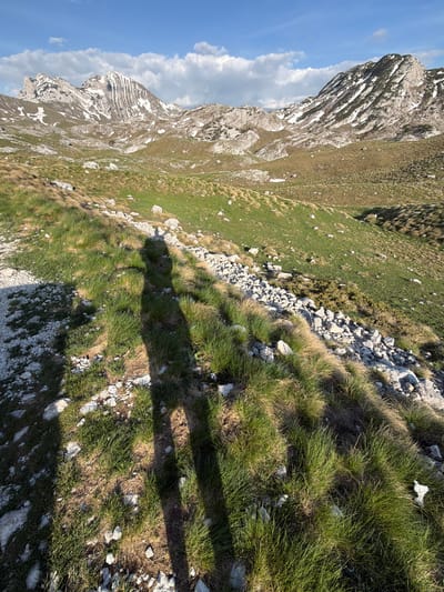 The image depicts a mountainous landscape near Boričje, Montenegro. In the foreground is grass interspersed with rocks. There is a shadow cast across the vegetation. To the left of the frame is a rocky mountain with layered rock formations and some snow patches. The sky is blue with some clouds visible.