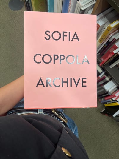 The image displays an overhead perspective of a person holding a book in an indoor setting. The central subject is a book with a solid pink cover, prominently displaying the title "SOFIA COPPOLA ARCHIVE" in capital letters. The word "SOFIA" is printed in black. The word "COPPOLA" features alternating black and silver-reflective characters: 'C' (black), 'O' (silver-reflective), 'P' (black), 'P' (black), 'O' (silver-reflective), 'L' (silver-reflective), 'A' (silver-reflective). The word "ARCHIVE" similarly alternates: 'A' (silver-reflective), 'R' (black), 'C' (silver-reflective), 'H' (black), 'I' (silver-reflective), 'V' (black), 'E' (silver-reflective).
A person's forearms are visible, holding the book from the lower left. Their lower torso, clad in blue denim jeans, is visible at the bottom of the frame, along with a dark belt featuring a rectangular gold-colored buckle. A dark-colored garment, possibly a jacket or blazer with circular gold-colored buttons, covers portions of their upper thighs.
The background features multiple tall bookshelves densely packed with books of various sizes and spine colors, indicating a bookstore or library environment. The flooring, visible on the left and bottom right, is a light-colored carpet, possibly beige or muted green. No specific visual cues or identifiers within the image provide confirmation of a precise location within El Paso, United States.