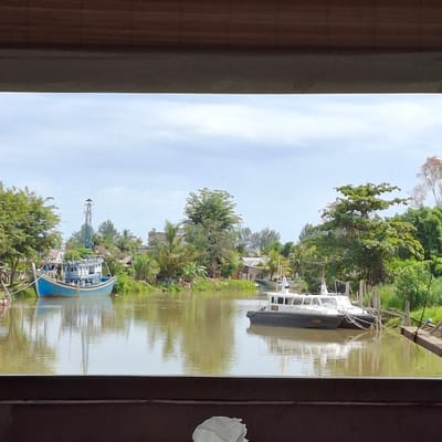 The image captures a serene canal scene in Mesjid Tuha, Indonesia. The canal, with its murky brown water, reflects the overcast sky and the lush green vegetation lining its banks. Two boats are the primary subjects. A blue and white fishing boat is moored to the left, its weathered exterior suggesting it is actively used. To the right, a sleek white and dark-hulled patrol boat is anchored, its presence hinting at security or enforcement operations. The background reveals a cluster of buildings, some appearing to be houses, interspersed with abundant tropical trees and palm fronds, characteristic of a coastal or riverside settlement. A tall, slender structure, possibly a lighthouse or a communication tower, stands behind the fishing boat, adding to the unique landscape. The foreground is framed by dark, horizontal wooden elements, suggesting the photo was taken from a covered vantage point or a structure overlooking the canal. No people are clearly visible in the image, contributing to the tranquil atmosphere.