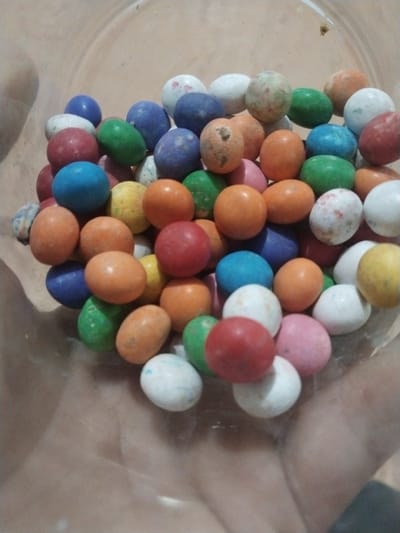 The image shows a close-up view of a collection of brightly colored, round candies, likely chocolate-covered peanuts or similar sweets. The candies are in various colors, including red, orange, blue, green, white, and yellow. They appear to be held within a transparent container, possibly a bowl or jar. The surface of the container is also visible, and there is no additional information about the surrounding environment. The location is provided as Keudekarieng, Indonesia, but the image itself gives no visual cues or context to verify this location. No people, actions, or interactions are depicted.