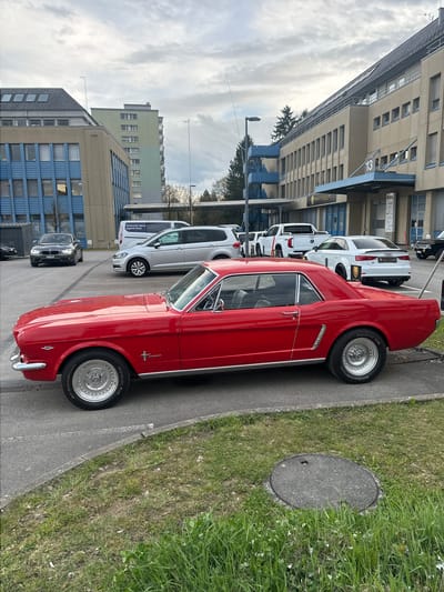 The image shows a red vintage Mustang parked on a paved area next to a sidewalk with grass. The car is facing forward, with its front end visible. In the background, a row of parked white vans and a dark-colored car are visible, parked next to a building. The building has a light beige exterior with multiple windows and the number "13" displayed prominently. "RIAG" can be seen next to 13. The building style appears to be commercial. The sky is overcast with varying cloud cover. The image appears to be taken in Wetzikon (ZH), Switzerland, as the white vans have German writing on them saying "Starte jetzt deine digitale Reise.". The word "VETTICE" is visible near a white car. A manhole cover is on the right.