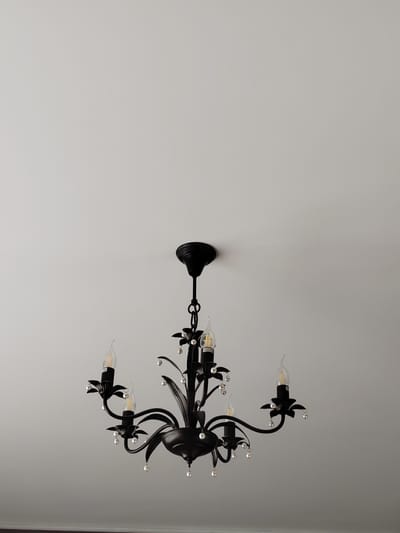 A black, five-light chandelier with faux-candle bulbs and clear crystal embellishments hangs from a white ceiling. The chandelier has ornate, curved arms and decorative floral elements at the base of each light. The ceiling appears to be a plain white surface, with no other visible features. The lighting in the image is soft and diffused, suggesting it may be daytime or the chandelier is not illuminated. The image is framed from a low angle, looking up at the chandelier. There are no people or other objects present in the scene. The location of the image is specified as Грачёвка, Russia.