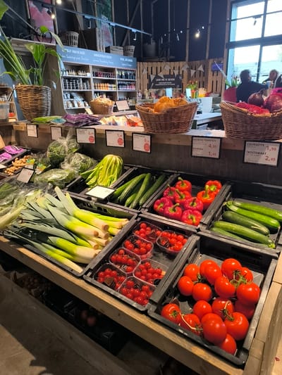 Here is a detailed description of the image:
**Scene:** An indoor produce market in Warmenhuizen, Netherlands.
**Subjects/Objects:**
* A variety of fresh vegetables are prominently displayed in black plastic crates on a wooden counter. These include:
* Leeks
* Cucumbers
* Red bell peppers
* Zucchinis (Courgettes)
* Cherry tomatoes
* Tomatoes on the vine
* Other vegetables (spinach, broccoli and onions) are displayed above
* Price tags indicating the cost of the produce.
* Behind the produce display is a shelf unit containing bottled goods, jams and preserves.
* Baskets containing what looks like bread and onions.
* There is a large leafy plant in a woven basket to the left.
* In the background, there is a window with daylight coming through, as well as people.
**People:**
* There appear to be two women standing together in the background near the window.
**Actions:**
* The produce is arranged for sale.
* Customers are shopping (implied by their presence in the background).
**Environment:**
* The market has a rustic feel with exposed wooden beams and shelving.
* The lighting is a mixture of natural daylight and artificial lights.