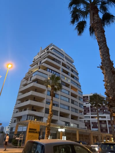 In Calp, Spain, a towering, modern, multi-story apartment building with many balconies and windows dominates the skyline. The building is light-colored and has a stepped, angular design. In the foreground and to the right, a large, mature palm tree with a textured trunk and fronds is visible, partially obscuring the building. To the left of the building, a streetlamp with a bright orange light is illuminating the scene, indicating it is dusk or early evening. Another, slightly shorter apartment building with white and maroon accents is visible behind the main building. Several cars are parked on the street in front of the buildings. A person in dark shorts and a light-colored t-shirt is standing on the sidewalk near the entrance of the main building, which has a sign with orange and yellow lettering. The sky is a clear, deep blue, typical of a twilight sky.