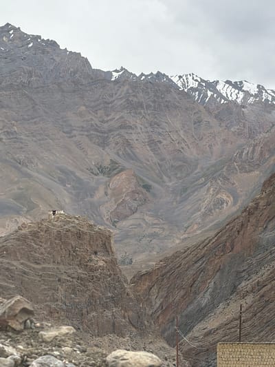 The image depicts a rugged, mountainous landscape with a small, white building perched on a rocky outcrop. The mountains are barren and rocky, with shades of brown and gray dominating the scene. Some of the higher peaks are capped with snow. The sky is overcast and a light gray. In the foreground, there are loose rocks and boulders. To the right of the frame, there are two wooden utility poles with wires and a brick wall. The building on the outcrop appears to be a monastery or a small temple. The overall impression is one of isolation and natural grandeur. The location is Zangla, India, a region known for its remote and beautiful mountainous terrain.