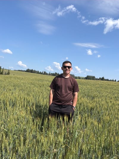 A young man with short dark hair, wearing sunglasses, a brown t-shirt, and dark shorts, stands in a field of tall green wheat under a blue sky with scattered white clouds. The man is positioned in the center of the frame, with his hands in his pockets. The wheat field extends into the background, where a line of trees and a gently sloping hill are visible. The scene appears to be in a rural, outdoor setting, possibly in the city of Podshivalovo, Russia, given the context. The lighting suggests it is daytime, with the sun casting shadows within the wheat stalks.
