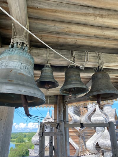 A close-up, low-angle shot shows four bronze bells hanging from a wooden structure, possibly a bell tower, in Boyarshchina, Russia. The largest bell, on the left, is a mottled greenish-bronze with ornate carvings around its rim and a rusty clapper hanging inside. Three smaller, darker bronze bells hang to its right, also with clappers. The bells are suspended by thick ropes and chains attached to weathered wooden beams and posts.
Behind the bells, through an opening in the wooden structure, a scenic view is visible. The background features a body of water, likely a lake or river, with lush green trees lining its banks. In the distance, a quaint village with distinctive wooden church domes, possibly onion domes, can be seen under a bright blue sky dotted with fluffy white clouds. The overall impression is one of a rural, historic setting, likely a church or monastery complex. The image captures the texture of the aged wood and metal, highlighting the craftsmanship of the bells and the traditional architecture of the area.