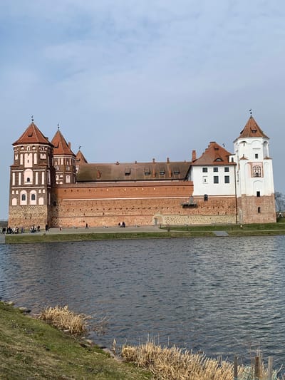 An outdoor, eye-level, wide shot captures the Mir Castle Complex situated within Мірскі сельскі Савет, Belarus, viewed across a body of water under a partially cloudy, light blue sky.
The prominent structure, Mir Castle, occupies the central and right portions of the image, presenting a facade composed primarily of red brick with white rendered sections and a rough-hewn stone base. The castle features multiple towers with reddish-brown tiled, conical roofs topped with finials. The left section displays several multi-story, octagonal towers with decorative white elements, arched window frames, and small rectangular windows. A long, lower brick wall connects these towers to a white-rendered building and a square tower on the far right, which also features a conical roof and a visible clock face with Roman numerals on one side. Small, dark rectangular windows are visible across the castle's various sections.
In front of the castle, a paved area separates it from the water. Approximately 20-30 individuals, depicted as indistinct figures, are observed walking or standing on this paved area and a connecting pathway near the castle's base. They appear to be visitors. One individual walks alone towards the right; others are in small groups.
The lower half of the frame is dominated by a dark blue-grey body of water, exhibiting small surface ripples. The immediate foreground on the left shows a sloped grassy bank with patches of dry, light brown reeds and some visible rocks at the waterline. Similar dry reeds are visible along the water's edge on the right. The horizon behind the castle appears clear.