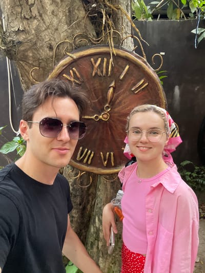 A vertical selfie captures a man and a woman positioned in front of a large, rustic clock face in an outdoor setting, likely a garden in Nam Nha Trang, Vietnam.
In the lower left, a man with dark hair wears dark sunglasses and a black t-shirt, looking towards the camera with a neutral expression. Behind him and slightly to the right, a smiling woman with blonde hair and clear-framed glasses looks directly at the camera. She is dressed in a pink t-shirt, a pink long-sleeved button-down shirt, and a red skirt with white polka dots. A colorful headscarf in shades of pink, yellow, black, and white is tied on her head. She holds a silver smartphone in her right hand, and a small flower pendant hangs from a necklace around her neck.
The prominent background feature is a large, ornate clock face crafted from wood, detailed with gold-colored Roman numerals (including I, II, IV, V, VI, VII, VIII, X, XI) and decorative hands. The clock is surrounded by golden metallic swirls and appears to be mounted on or intertwined with a textured, tree-like structure. Green foliage is visible in the background, contrasting with darker elements. The scene is brightly lit, indicating daytime.