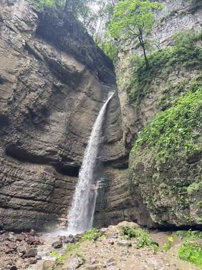 The image depicts a waterfall in a canyon, likely part of the Nizhniy Chegem region of Russia. The waterfall cascades down a sheer, rocky cliff face that is covered in sparse vegetation in places. The water falls into a shallow, rocky stream at the bottom of the canyon. The surrounding canyon walls are steep and composed of layered rock formations. The overall impression is one of a wild, natural landscape. There are no people or animals visible in the image. The scene is bathed in natural daylight, with a slightly overcast sky suggested by the diffuse lighting. The environment is damp and rocky, with evidence of the water flow at the base of the waterfall.