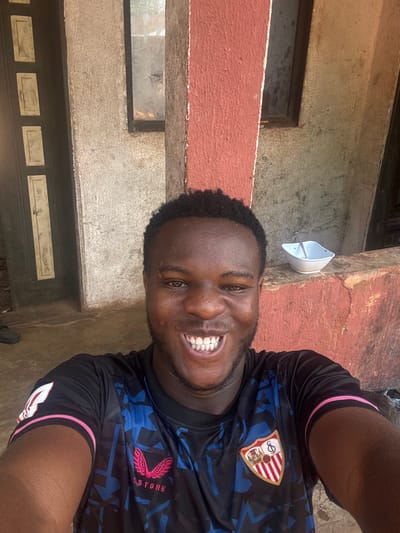 The image depicts a smiling dark-skinned male individual taking a selfie in Aguata, Nigeria. He has short, dark, curly hair, a wide smile with visible teeth, and a faint scar above his left eyebrow. He is wearing a black and blue short-sleeved jersey with a Sevilla FC crest on the right chest, a Castore logo on the left chest, and pink trim on the sleeves. The background consists of an outdoor or semi-outdoor area featuring a prominent reddish-brown painted pillar. To the left of the pillar, a dark door with two light-colored vertical decorative panels is partially visible. To the right of the pillar, a textured light grey wall with two recessed rectangular window openings is present. A white bowl containing a single utensil rests on a ledge to the right of the subject's head. The scene is illuminated by daylight.