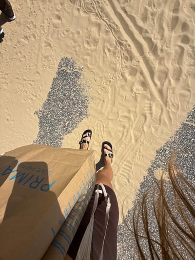 The image depicts a person's legs and feet standing on a sandy area, possibly a beach or promenade, in Bournemouth, United Kingdom. The sand is marked with numerous footprints. To the right of the person, there is a section of pebbled ground. The person is wearing dark sandals and holding a brown paper shopping bag with blue lettering that appears to read "PRIMARK LOVE THE FEEL". The person's hair is long and brown and is visible in the bottom right of the frame, suggesting it is blowing in the wind. The lighting indicates it is daytime and sunny, casting shadows from the person and the shopping bag. Another person's legs and feet, wearing white sneakers, are partially visible in the upper left corner, also standing on the sandy ground. The context of Bournemouth, UK, combined with the presence of a Primark bag, suggests a shopping excursion in a coastal area.