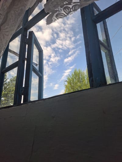 An open window frames a view of a blue sky with scattered white clouds. Outside the window, green trees and utility wires are visible. The window frame is made of weathered blue wood, and part of a white lace curtain is visible at the top left. The interior wall below the window is textured and painted a light color. The scene is likely from Sayhun, Uzbekistan.
