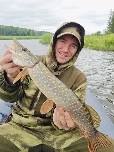 A man in a camouflage jacket and hat smiles at the camera while holding a pike in a boat. The man is in the foreground, with the pike held horizontally across his chest. The background shows a body of water, with trees and reeds on the far bank. The sky is overcast. The man appears to be on a fishing trip in the village of Detskii Dom (Bashenino), Russia.