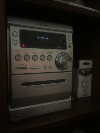 The image shows a close-up view of a silver micro hi-fi component system, likely a stereo, with its brand name "AWA" visible at the top. The system has a digital display, various buttons for functions like "DISPLAY," "TAPE," "CD," "TUNER/BAND," "PLAY MODE/TUNING MODE," "TUNING +/-," "- ALBUM +," "CD SYNCRO," and "REC." There is also a large volume knob. A red power indicator light is illuminated on the front panel. Below the main control panel, there is a CD player slot with the text "FULL LOGIC OPERATION" printed below it. The system is placed on top of another component, and a small box with text "BULBO LED 7W" is visible to the right of the stereo system, suggesting it is on a shelf or in a storage unit. The overall environment appears to be indoors, likely a home or a store, with dark shelving. The image does not contain any information that can be used to identify its location in San Antonio, Venezuela.