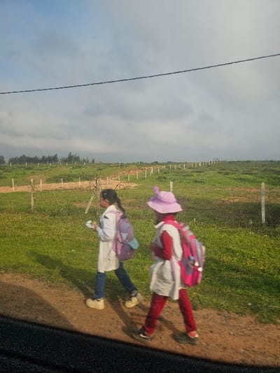 Two young females, appearing to be students, are walking right-to-left along a dirt path in a rural area of Ameur عامر, Morocco. The subject on the left wears a white smock over blue jeans and light-colored shoes, carrying a purple backpack and holding a small white object in her right hand. The subject on the right, slightly behind, wears a white smock over red trousers and dark shoes, a light purple wide-brimmed hat with two protrusions, and a pink and purple backpack. The environment consists of a green grassy field on either side of the path, marked by a line of white fence posts extending into the distance. A treeline is visible on the far-left horizon. The sky is overcast with a mix of light blue and grey clouds, and a dark utility line stretches horizontally across the upper frame. Shadows indicate daylight from the left.