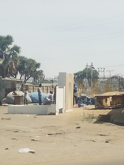 An outdoor scene in Darmanawa, Nigeria, depicts a dusty, unpaved open area under a clear sky. In the mid-ground, a metal fence separates the foreground activity from a line of palm trees and utility poles with power lines in the background.
In the left mid-ground, a corrugated metal structure is partially visible behind several large, irregularly shaped bundles covered in light blue and grey material. Adjacent to these bundles, a wooden table is present. Two individuals, appearing to be young males, are near this table; one stands beside it, while another stands further left near the bundles.
Centrally, a partially assembled or dismantled light-colored modular structure, consisting of white and beige panels, stands on a raised platform. To the right of this structure, several more individuals are present; one male in a light shirt and dark trousers stands near the structure, and others are partially obscured behind it, with one appearing to be squatting.
On the right side of the mid-ground, a large, rusty, open-topped industrial container is visible. A person in a blue top and light-colored trousers stands near this container. Two blue, wheeled air compressors or similar machinery are positioned on the ground in front of the container.
The foreground consists of barren, light brown earth with scattered debris, including a white plastic bag. The overall impression is that of an informal outdoor workspace or gathering area.