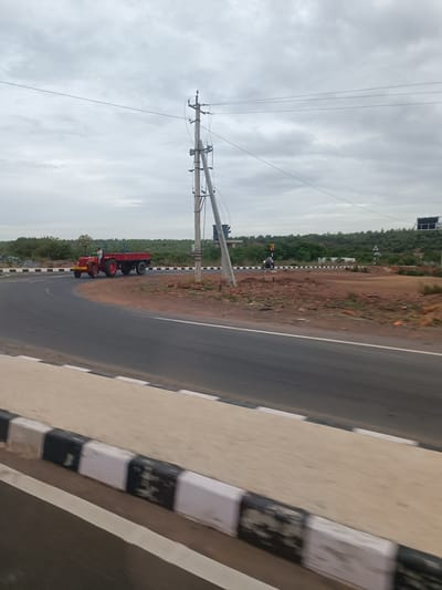 The image shows a rural road scene near Betamcherla, India. A red tractor with a trailer is driving on the road. A person is visible on the tractor. The road is paved and curves to the left. There is a black and white striped concrete barrier along the side of the road. In the background, there are trees and hills, with a cloudy sky overhead. A utility pole with wires stands near the road. Another vehicle, possibly a motorcycle, is also visible on the road in the distance. The terrain appears dry and rocky.