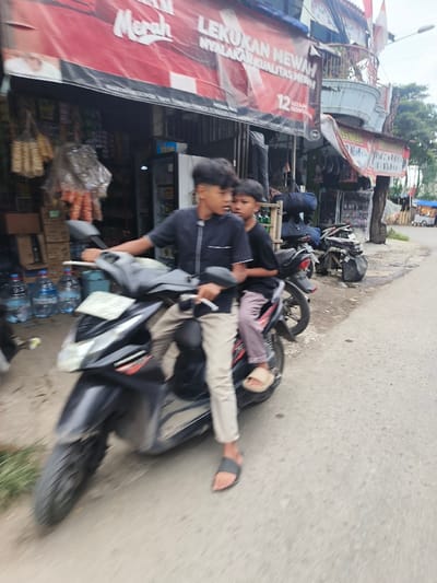 Two boys ride a black motorbike on a paved road next to a building. The rider wears a navy shirt, tan pants, and flip-flops. The passenger has on a black shirt, maroon pants, and slides. Bottles of water sit stacked beside the storefront, which is covered by a red banner. Other motorbikes are parked further down the road. The image appears slightly blurry.