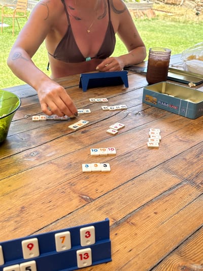 A close-up, eye-level shot shows a person's hands playing a game of Rummikub on a weathered, wooden picnic table outdoors. The player, identified by their hands with patterned fingernails and arm with tattoos, is in the process of placing tiles on the table. Some of the visible tiles are marked with numbers like "3," "10," "12," and "13." There are two blue Rummikub tile racks, one in the foreground containing tiles with the numbers "9," "7," "3," and "13," and another out of focus in the background, holding a tablet. A green glass bowl filled with what appears to be popcorn is situated to the left of the frame, and a glass filled with a dark beverage, ice, and a straw is to the right, next to a partially opened Rummikub box. The setting is outdoors, with blurred green foliage and part of a rustic chair visible in the background, suggesting a casual, possibly garden or patio environment. The lighting suggests it is daytime. The scene does not provide any specific landmarks or indicators to confirm a location in San Lorenzo De Tormes, Spain.