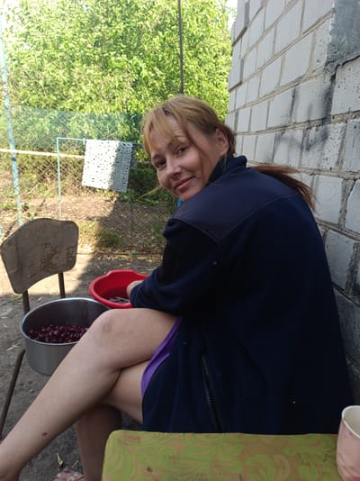 An outdoor, medium shot shows a middle-aged woman with auburn hair and fair skin sitting on a chair next to a brick wall. She is wearing a dark blue jacket and a purple garment underneath. Her legs are crossed, and she is looking towards the camera with a slight smile. In front of her are two bowls: a silver metal bowl filled with red cherries and a red plastic bowl containing water and cherries. Behind the woman, a chain-link fence and lush green trees are visible. The scene appears to be in a backyard or garden area, possibly in the city of Saksahan, Ukraine. The lighting suggests it is daytime, with sunlight casting shadows on the wall.