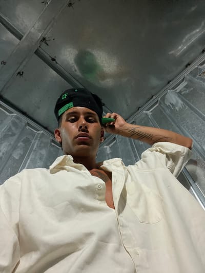 A man in a light-colored shirt and black baseball cap stands inside a metallic structure. He holds a green object near his ear. He has tattoos on his neck and arm. The walls and ceiling around him are metallic, possibly a truck bed or some kind of container. The photo is taken from a low angle, looking up at the man.