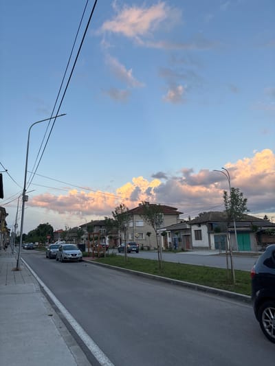 This image captures a street scene in Popovo, Bulgaria, during the late afternoon or early evening, characterized by a vibrant sky. The perspective is from the left side of an asphalt road, looking forward along its length.

The upper two-thirds of the frame are dominated by the sky, which transitions from a clear, light blue overhead to a dramatic display of clouds along the horizon. These cumulus clouds exhibit prominent pink, orange, and white hues, strongly illuminated by the low-angle sun, creating a striking contrast with the cooler blue sky. A faint, slender crescent moon is visible in the upper right portion of the blue sky.

On the immediate left, a concrete sidewalk runs parallel to the asphalt road, separated by a distinct white curb line. A tall, grey metal streetlight pole with two horizontal luminaires extends from the left, with multiple dark utility lines strung across the upper-left quadrant of the frame. Further down the sidewalk, indistinct figures of several pedestrians can be seen. Buildings lining the left side of the street appear to be multi-story residential structures, some with visible balconies and varied architectural details. A red circular "No Entry" sign is visible in the mid-distance on this side.

The asphalt road surface appears to be in fair condition. Several vehicles are parked along the left curb. These include a dark grey sedan, a silver SEAT Ibiza bearing Bulgarian license plate "T 4472 AX," and another dark sedan further behind it.

On the right side of the street, a narrow strip of maintained grass separates the roadway from the adjacent properties. Several young, slender trees, supported by light-colored wooden frames, are planted within this grassy strip. Another grey metal streetlight pole, identical in design to the one on the left, is visible further down this side. The buildings on the right consist of a mix of older, single-story structures with tiled roofs and one more modern, two-story building with a light-colored facade. One older building features prominent green garage doors or shutters. A yellow diamond-shaped road sign, indicating a priority road, is mounted on a pole near one of the buildings. The rear quarter of a dark-colored vehicle, likely an SUV, with a multi-spoke alloy wheel, is partially visible in the extreme lower right corner, parked facing away from the viewer.

The scene conveys a tranquil, residential urban environment under the early evening sky.
