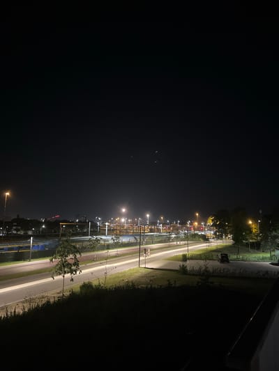 The image depicts a nighttime scene in Venlo, Netherlands, likely near a train station or transportation hub. A train with blue and yellow cars is visible on tracks. A road with cars is illuminated by streetlights, creating light trails due to a long exposure. The foreground features dark foliage and a concrete barrier. In the background, more streetlights, buildings, and trees can be observed against a dark sky. The overall atmosphere is that of an urban area at night.