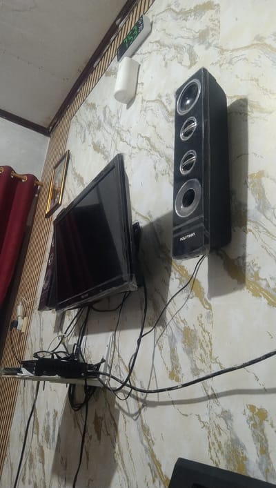 The image depicts a wall in what appears to be a room in Reudeuep, Indonesia. The wall has a marble-like pattern with gold and gray veining. Mounted on the wall are a black flat-screen television with a plastic protective cover, and a tall, black speaker with three circular elements. Above the television and speaker, a digital clock displaying "7:53" and "28" is mounted. A white cylindrical object is mounted below the clock. A small framed picture with a gold frame is visible to the left of the television. Red curtains are partially visible on the far left. A white shelf is mounted below the television, laden with various black electronic devices and a tangled mass of black cables. The lighting suggests an indoor environment.