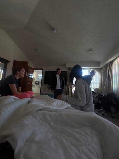 The image depicts an interior scene, identified as a bedroom in Cacalomacan, Mexico. Four individuals are present under daylight conditions, with natural light entering from a window on the right. In the foreground, a bed with white, rumpled bedding is visible. From left to right: A female, with brown hair, wears a dark short-sleeved top and pink trousers. She is seated, smiling, and oriented towards the center-right. Standing centrally is a male in a dark puffer jacket over a white shirt and dark trousers; he is looking towards the right. To his right, a second female is seated with her back mostly to the camera, her dark hair obscuring her face from this perspective. She wears a light grey long-sleeved top and light grey trousers. Partially visible behind the second female, on the far right, is a second male wearing a light-colored baseball cap and a dark top; he is positioned as if leaning or peeking from behind the second female and looking towards the camera. The background includes a large, wall-mounted flat-screen television. To the left, a dark wooden doorframe leads into an adjacent, brightly lit room. To the right, floor-to-ceiling curtains cover a window, and a desk chair with a dark object draped over it is positioned near a partially visible desk. The light-colored ceiling features an angled segment and two circular fixtures, likely smoke detectors.