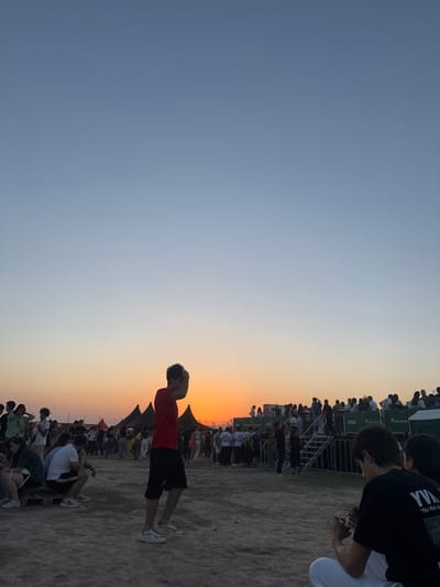 This image depicts an outdoor event at sunset in Yeghvard, Armenia. The sky transitions from a clear, light blue at the zenith to a vibrant orange and yellow at the horizon, where the sun is setting. The foreground consists of dry, light-colored ground, possibly dirt or gravel.
In the left-center foreground, a person wearing a red short-sleeved shirt, black shorts, and light-colored athletic shoes is in motion, with both hands raised near their head. To the right in the foreground, two individuals are seated on the ground, with one visible from the back, wearing a black t-shirt with white text reading "YWA" and partially visible "The Pick Go...".
The midground is densely populated with a crowd of people, some standing and others seated on wooden pallets. Several dark, pointed tent structures are visible among the crowd. In the right midground and background, an elevated, multi-level metal structure with stairs and railings is crowded with additional attendees. Green banners displaying "VISA" and another logo (partially visible, resembling "ARDSHINBANK") are affixed to this structure, indicating sponsorship. The overall scene suggests a festival or public gathering during twilight.