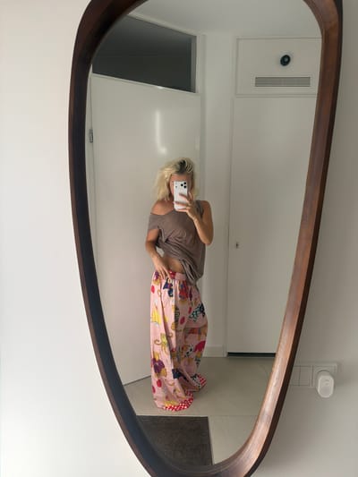 A blonde woman, reflected in a large, oval, dark wood-framed mirror, stands in a well-lit, modern interior. She is wearing a taupe, off-the-shoulder top and pink, wide-leg pajama pants adorned with a colorful pattern of leopards, moons, and other whimsical figures. Her hair is styled in a messy bun. She holds a smartphone in her right hand, taking a selfie, and her left hand rests on her hip. The background of the reflection reveals a white wall with a closed door, a ventilation grille, and a white electrical outlet with a small white device plugged into it. The floor appears to be tiled with beige tiles. The overall impression is one of a casual, domestic setting. This image was captured in a home in Ridderkerk, Netherlands.