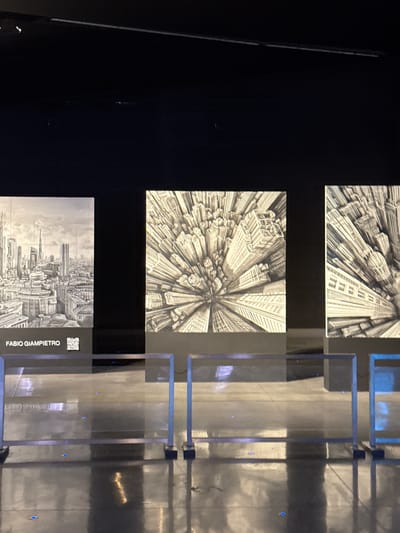 The image depicts an art exhibition featuring three large black and white drawings of cityscapes, likely rendered in pencil or ink. The drawings are displayed on dark pedestals against a black background. The leftmost drawing shows a wide cityscape with numerous buildings of varying heights, including skyscrapers. Below this drawing, the name "FABIO GIAMPIETRO" is visible, along with a QR code. The other two drawings present abstract, distorted perspectives of city buildings, with a strong sense of converging lines and depth, appearing to zoom outwards from a central point. These drawings are also presented on dark pedestals.
In the foreground, there are metal stanchions with horizontal bars, creating a barrier that separates the viewer from the artwork. The floor is highly reflective, showing the reflections of the stanchions and some scattered blue lights. The overall lighting is dim, with spotlights illuminating the artwork and creating reflections on the floor. The scene suggests an indoor exhibition space.
Based on the provided information, this exhibition is located at the Studio Museo in Ferno, Italy.