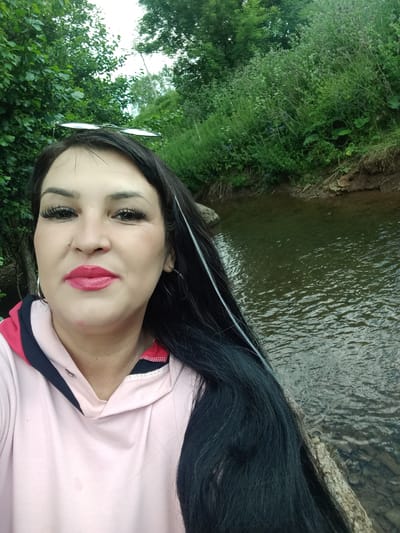 The media file is a selfie of a woman with long, dark hair and bright red lipstick. She is wearing a pink hoodie with navy blue and red accents. She is looking at the camera with her head tilted. Behind her is a body of water with a rocky edge, and lush green trees and vegetation. There are no visible text or specific details about the time of day or weather, but the lighting suggests it is daytime. The overall mood seems relaxed and casual. The setting appears to be a natural outdoor environment, possibly a park or a riverbank.