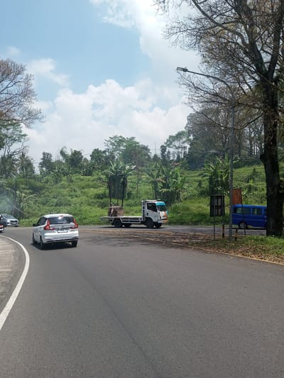 The image shows a road scene with vehicles in what appears to be a rural area. A white car is driving on the left side of the road. A flatbed truck and a blue van are visible in the distance on the right side of the road. The background consists of lush green vegetation and trees. A street light is positioned near the blue van. The sky is partly cloudy. There is no clear indication of Nawng Hkar, Myanmar.