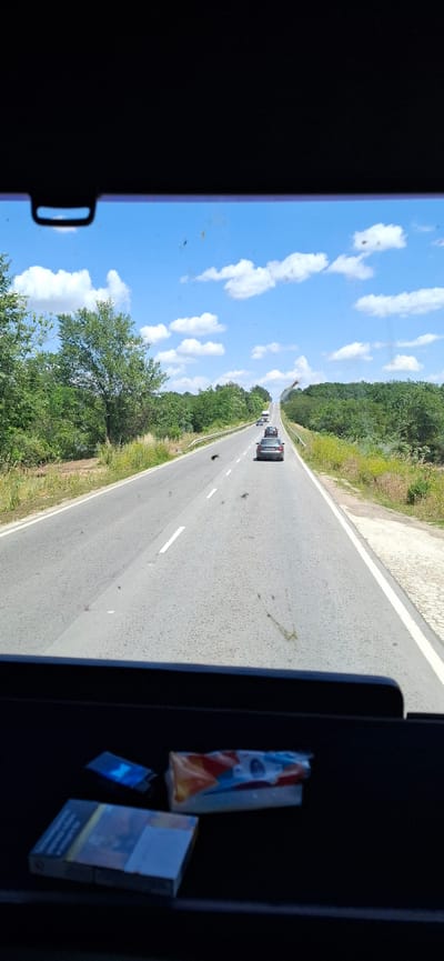 The image shows a view from inside a vehicle, looking out onto a highway. The perspective is tilted, giving a sense of motion. The sky is bright blue with scattered white clouds, suggesting a clear, sunny day. Green trees line both sides of the road, with some dry grass and vegetation visible along the shoulder. Several cars are driving on the highway, appearing as dark shapes in the distance.
In the foreground, on the right side of the frame, there are several objects inside the vehicle. These include a pack of cigarettes and a lighter. The pack of cigarettes displays colorful branding with text, though it is not clearly legible. The lighter appears to be blue. The interior of the vehicle is dark, creating a contrast with the bright outdoor scene.
There is no clear indication of people or specific emotional states. The overall impression is that of a casual drive through a rural or semi-rural area. The presence of the cigarettes and lighter suggests the driver or a passenger might be a smoker. The time of day appears to be daytime, given the bright sunlight and blue sky. The location cues of trees and a highway suggest an outdoor, travel-related context. The overall scene is somewhat cluttered with dirt or debris on the window, which is partially visible on the left side of the frame.