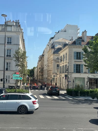 The image depicts a street scene in Neuilly-sur-Seine, France, with a view looking down a tree-lined avenue. On the left, a white apartment building with grey mansard roofs and numerous windows is visible. A street lamp with a circular fixture stands in front of this building. Green street signs indicating "N 13", "LA DÉFENSE", and "NEUILLY-CENTRE" are mounted on a pole.

The right side of the street features a row of cream-colored buildings with light blue shutters, typical of Parisian architecture. These buildings have multiple stories and ornate facades. One building has a shop with a large glass storefront labeled "CYRILLUS".

In the foreground, a white station wagon is driving on the asphalt road. Further down the street, a black SUV is also in motion. There is a crosswalk marked on the road, and temporary orange barriers with reflective tape suggest ongoing construction or roadwork. A few pedestrians are present, walking on the sidewalks or crossing the street. The sky is clear and blue. Reflections are visible on the surface from which the photograph was taken, likely a window.